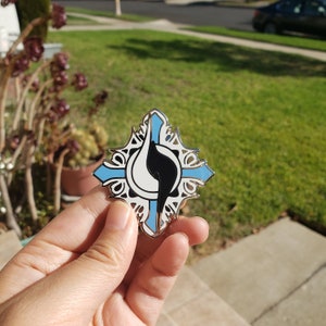 Puede incluir: Un pin de esmalte plateado y azul con un símbolo yin yang blanco y negro en el centro. El pin tiene forma de diamante con intrincados detalles alrededor de los bordes.