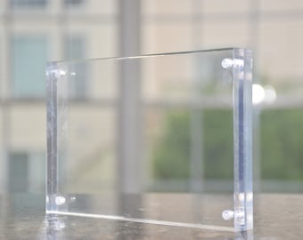 May include: A clear acrylic picture frame with rounded corners and silver metal clips on each side. The frame is standing on a dark surface.