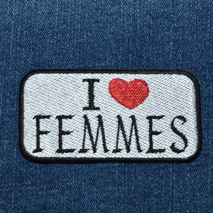 May include: A white patch with black trim on a blue denim background. The patch has the text "I love Femmes" with a red heart in between the words.