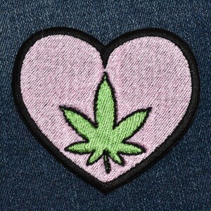 May include: A pink heart-shaped patch with a green marijuana leaf embroidered in the center. The patch has a black outline.