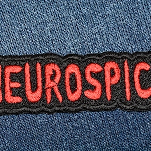 May include: Embroidered patch with the word "NEUROSPICY" in red letters on a black background. The patch has a black border and is set against a blue denim fabric.