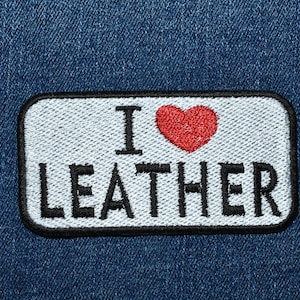 May include: A white patch with black stitching on a blue denim background. The patch has a red heart and the text "I LOVE LEATHER" in black.