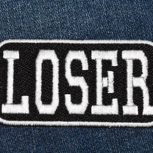 May include: A black and white embroidered patch with the word "LOSER" in white letters on a black background. The patch is sewn onto a blue denim fabric.