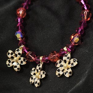 Peut inclure: Un bracelet de perles roses et oranges. Il comporte trois breloques en forme de fleurs dorées avec un motif à carreaux noir et blanc et des accents de cristal. Le bracelet est présenté sur un fond noir.