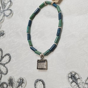 Puede incluir: Un brazalete de cuentas turquesa y azul con un dije de plata en forma de una pequeña caja. El brazalete está hecho a mano y tiene una etiqueta que dice "Leslie's Stroke Designs Handmade Crafts".