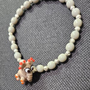 May include: A gray beaded bracelet with a silver charm and a glass elephant charm with red and orange accents.