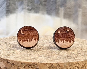May include: A pair of wooden stud earrings with a nature theme. One earring features a crescent moon over a forest, the other has stars. The earrings are round and sit on a cork surface.