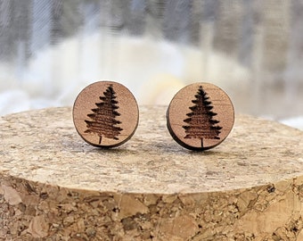 May include: A pair of wooden stud earrings. Each earring is a small, round, light brown disc with a detailed, etched image of a pine tree. The earrings are displayed on a cork surface, with a blurred background.