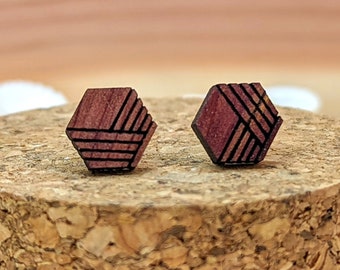 May include: A pair of geometric wooden stud earrings. Each earring is a reddish-brown hexagon with a black linear design. The earrings are displayed on a cork surface, surrounded by white seashells.