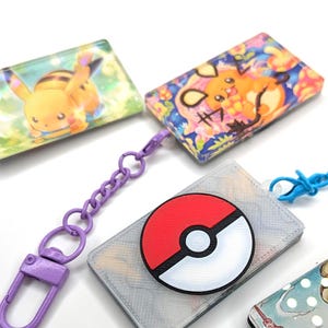 May include: Four rectangular badge holders with attached plastic chains and clasps. One features a red and white Poké Ball emblem, while the others display colorful cartoon character illustrations including a yellow creature and a brown creature.