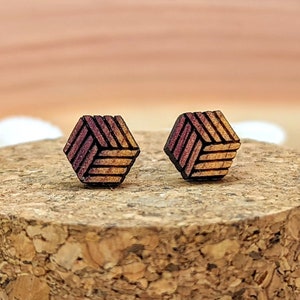 May include: Two wooden stud earrings with a geometric cube design. The earrings feature a pattern of intersecting lines in shades of red and gold. They are displayed on a cork surface surrounded by seashells, creating a natural aesthetic.