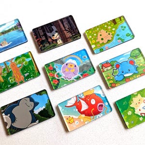 May include: A collection of rectangular, colorful magnets featuring various cartoon characters and scenic backgrounds. The magnets showcase a variety of designs, including landscapes, creatures, and whimsical illustrations. The magnets are arranged on a white surface.