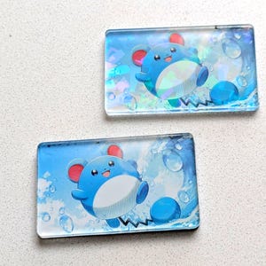 May include: Two rectangular, transparent acrylic charms featuring a blue cartoon character with red ears and a white belly. The background is a blue gradient with water droplet and wave designs. The character is smiling and appears to be swimming.