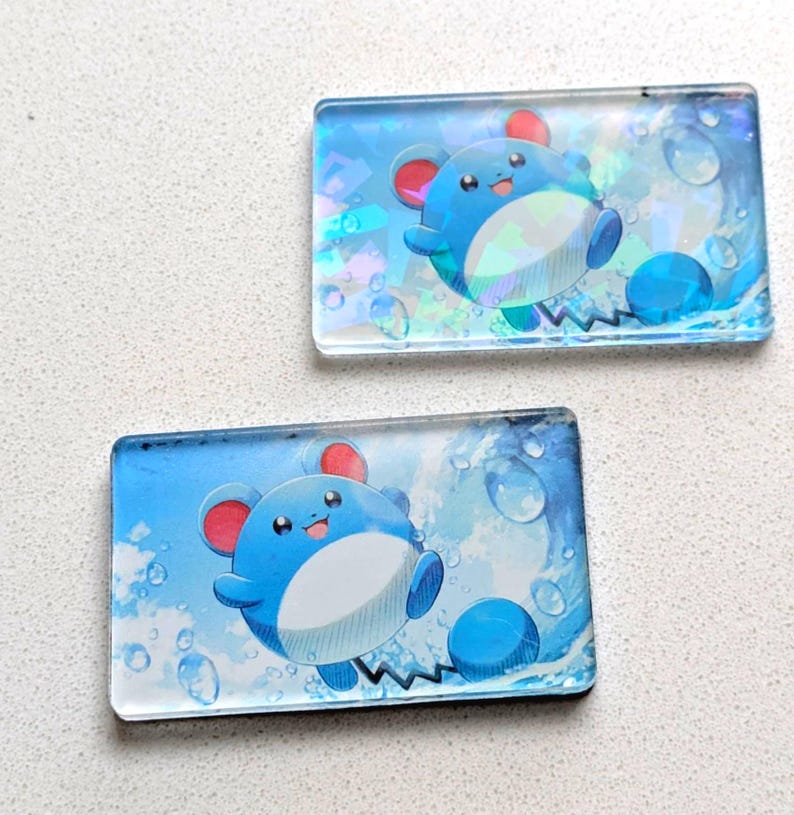 May include: Two rectangular, transparent acrylic charms featuring a blue cartoon character with red ears and a white belly. The background is a blue gradient with water droplet and wave designs. The character is smiling and appears to be swimming.