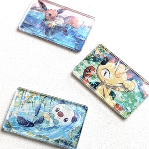 May include: Three rectangular, clear acrylic charms featuring colorful illustrations of Pok&eacute;mon characters. One charm depicts Eevee in a watery scene, another Meowth, and the third, Oshawott. The charms have a glossy finish.