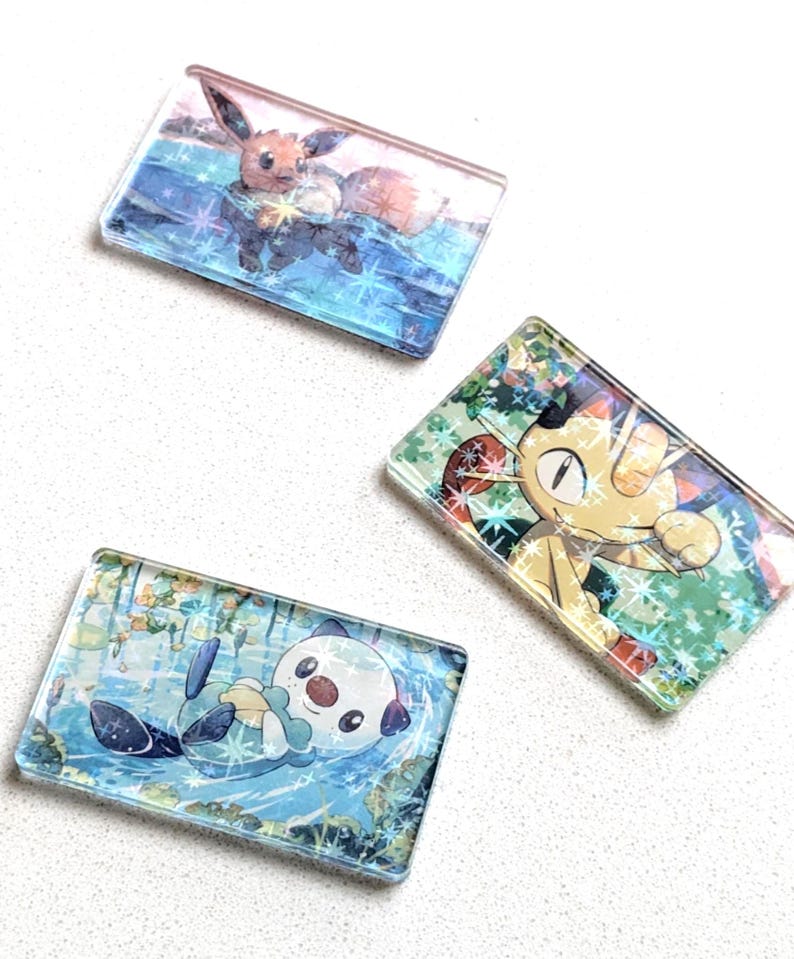 May include: Three rectangular, clear acrylic charms featuring colorful illustrations of Pok&eacute;mon characters. One charm depicts Eevee in a watery scene, another Meowth, and the third, Oshawott. The charms have a glossy finish.