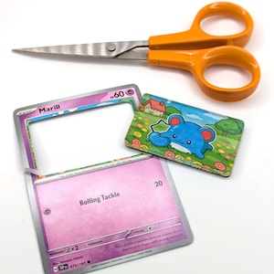 May include: An orange-handled pair of scissors rests above two Pok&eacute;mon trading cards and a small, rectangular illustration of a blue creature. One card has the name "Marill" and the text "Rolling Tackle". The illustration shows a blue creature in a field.