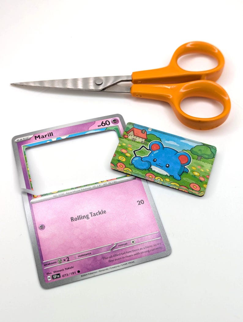 May include: An orange-handled pair of scissors rests above two Pok&eacute;mon trading cards and a small, rectangular illustration of a blue creature. One card has the name "Marill" and the text "Rolling Tackle". The illustration shows a blue creature in a field.