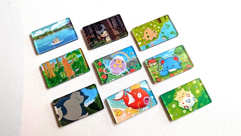 May include: A collection of rectangular, colorful magnets featuring various cartoon characters and scenic backgrounds. The magnets showcase a variety of designs, including landscapes, creatures, and whimsical illustrations. The magnets are arranged on a white surface.