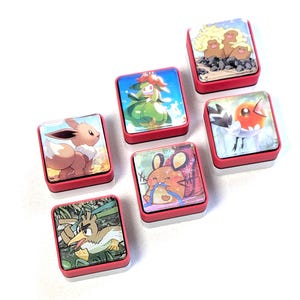 May include: Six square pins with red and white bases, each featuring a different colorful illustration of a cartoon creature. The designs include a brown fox-like character, a green plant-like figure, a bird, a rodent, and a group of three brown figures.