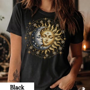 Puede incluir: Una camiseta negra de manga corta con un gráfico central de un sol dorado y una luna creciente plateada con rostros humanos, rodeados por una disposición circular de estrellas doradas y líneas finas.