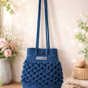 Puede incluir: Un bolso tote de ganchillo azul hecho a mano con asas largas. El bolso presenta un dise&ntilde;o festoneado texturizado en la parte inferior y un peque&ntilde;o adorno decorativo cerca de la parte superior. El bolso se exhibe sobre una superficie de madera.