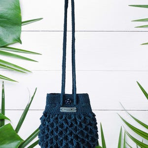 Puede incluir: Un bolso de ganchillo azul oscuro con una asa larga. El bolso tiene un dise&ntilde;o texturizado de tejido abierto en la parte inferior y una peque&ntilde;a etiqueta. El bolso cuelga sobre un fondo blanco con hojas verdes.