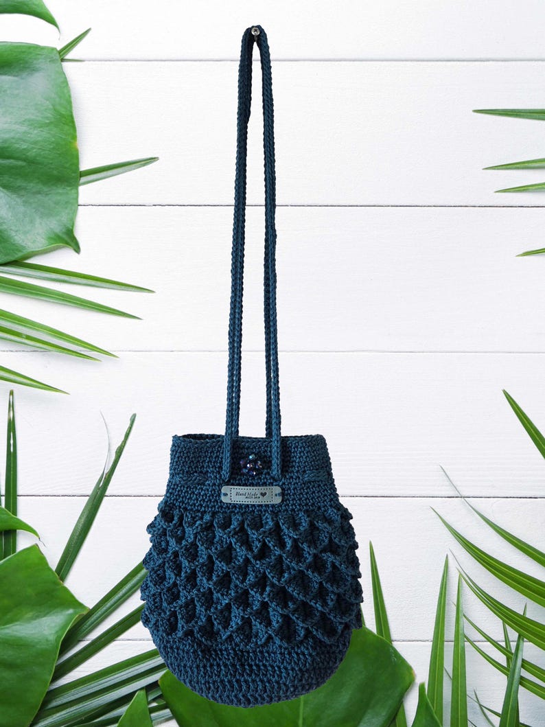 Puede incluir: Un bolso de ganchillo azul oscuro con una asa larga. El bolso tiene un dise&ntilde;o texturizado de tejido abierto en la parte inferior y una peque&ntilde;a etiqueta. El bolso cuelga sobre un fondo blanco con hojas verdes.