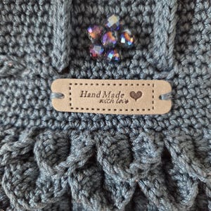 Puede incluir: Primer plano de un bolso de ganchillo azul hecho a mano con un patr&oacute;n texturizado. El bolso presenta un grupo decorativo de cuentas iridiscentes y una peque&ntilde;a etiqueta que dice "Hand Made with love" con un s&iacute;mbolo de coraz&oacute;n.