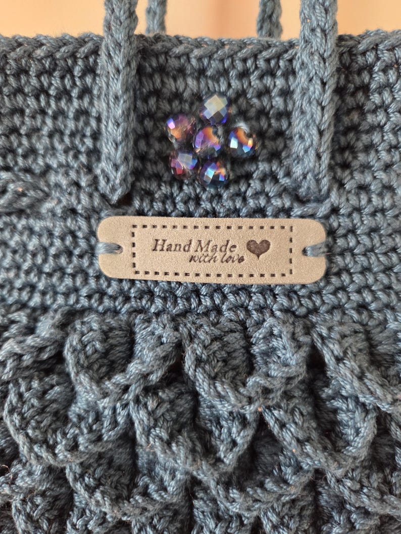 Puede incluir: Primer plano de un bolso de ganchillo azul hecho a mano con un patr&oacute;n texturizado. El bolso presenta un grupo decorativo de cuentas iridiscentes y una peque&ntilde;a etiqueta que dice "Hand Made with love" con un s&iacute;mbolo de coraz&oacute;n.