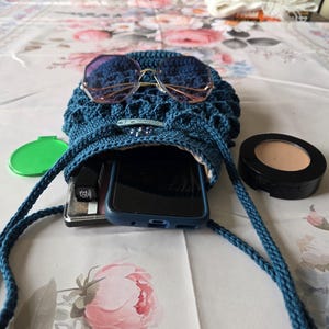 Puede incluir: Un bolso cruzado azul tejido a ganchillo con cierre de cord&oacute;n, que contiene un smartphone, maquillaje y otros accesorios. El bolso tiene gafas de sol encima. Tambi&eacute;n se ve un estuche verde y un estuche de maquillaje.