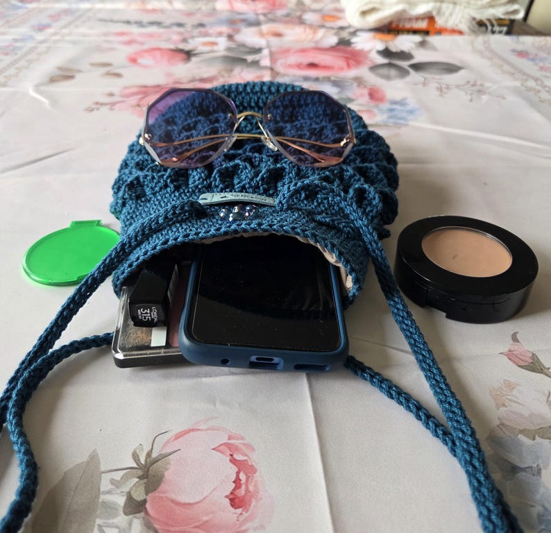 Puede incluir: Un bolso cruzado azul tejido a ganchillo con cierre de cord&oacute;n, que contiene un smartphone, maquillaje y otros accesorios. El bolso tiene gafas de sol encima. Tambi&eacute;n se ve un estuche verde y un estuche de maquillaje.