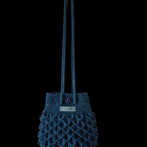 Puede incluir: Bolso de ganchillo azul oscuro hecho a mano con un patr&oacute;n texturizado en forma de escamas. El bolso tiene un cierre de cord&oacute;n y una correa larga. Una peque&ntilde;a etiqueta con texto est&aacute; adherida a la parte superior. El bolso est&aacute; suspendido sobre un fondo negro.