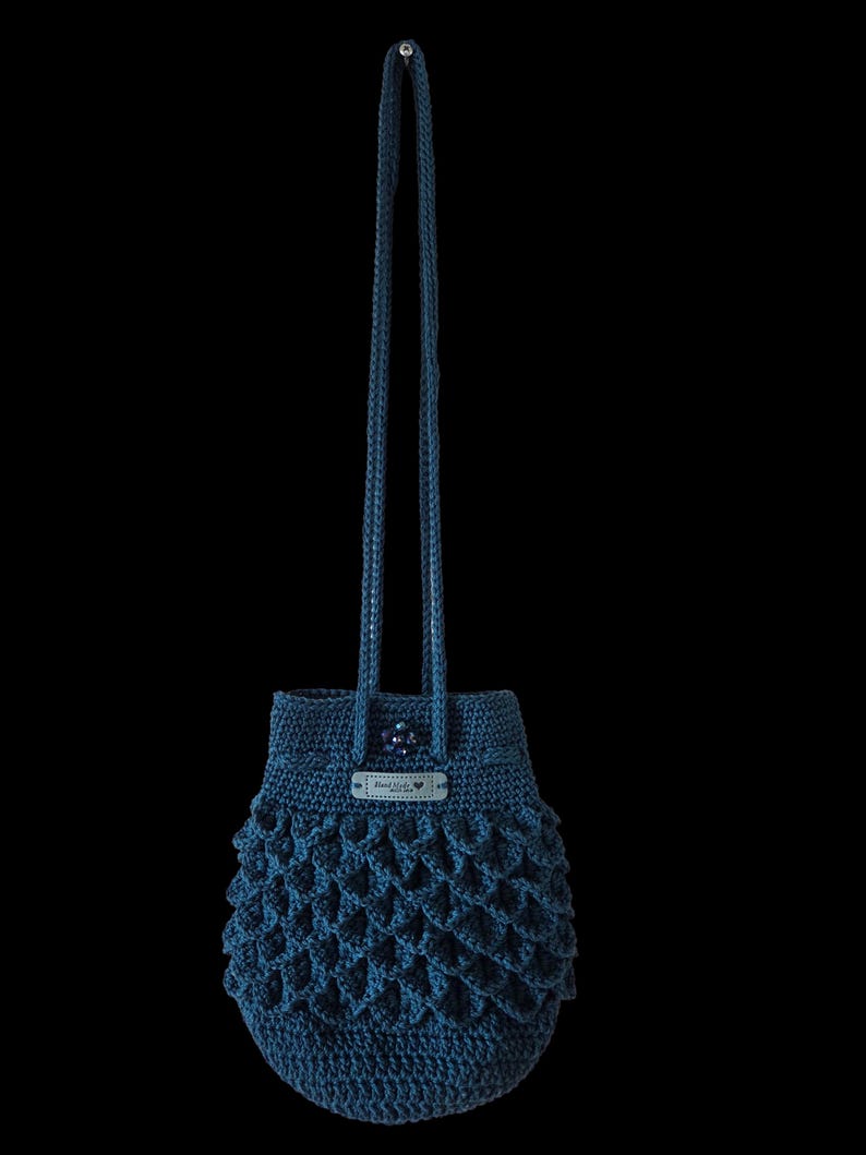 Puede incluir: Bolso de ganchillo azul oscuro hecho a mano con un patr&oacute;n texturizado en forma de escamas. El bolso tiene un cierre de cord&oacute;n y una correa larga. Una peque&ntilde;a etiqueta con texto est&aacute; adherida a la parte superior. El bolso est&aacute; suspendido sobre un fondo negro.