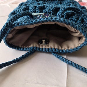 Puede incluir: Un bolso con cord&oacute;n de ganchillo azul turquesa con un patr&oacute;n texturizado. El bolso tiene un forro de tela beige y un cierre de bot&oacute;n a presi&oacute;n plateado. Cuentas decorativas est&aacute;n unidas a la parte delantera del bolso.