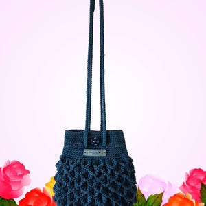Puede incluir: Un bolso de ganchillo azul oscuro con una correa larga. El bolso tiene un dise&ntilde;o texturizado de tejido abierto en la parte inferior y una peque&ntilde;a etiqueta que dice "Hand Made". El fondo es un degradado de rosa con rosas coloridas.