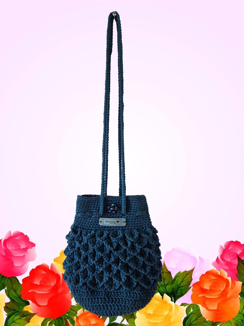 Puede incluir: Un bolso de ganchillo azul oscuro con una correa larga. El bolso tiene un dise&ntilde;o texturizado de tejido abierto en la parte inferior y una peque&ntilde;a etiqueta que dice "Hand Made". El fondo es un degradado de rosa con rosas coloridas.