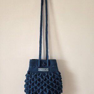 Puede incluir: Bolso de ganchillo azul oscuro hecho a mano con un patr&oacute;n texturizado en forma de escamas. El bolso tiene una correa larga trenzada y una peque&ntilde;a etiqueta con texto. El bolso cuelga contra una pared neutra.