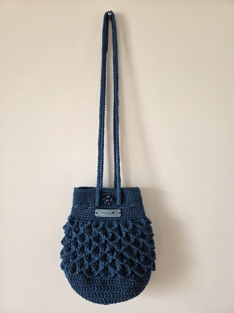 Puede incluir: Bolso de ganchillo azul oscuro hecho a mano con un patr&oacute;n texturizado en forma de escamas. El bolso tiene una correa larga trenzada y una peque&ntilde;a etiqueta con texto. El bolso cuelga contra una pared neutra.