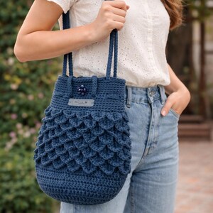 Puede incluir: Bolso de crochet azul marino hecho a mano con un patr&oacute;n &uacute;nico en forma de escamas. El bolso tiene un cierre de cord&oacute;n y correas resistentes. Una peque&ntilde;a etiqueta con texto y un bot&oacute;n decorativo a&ntilde;aden detalles.