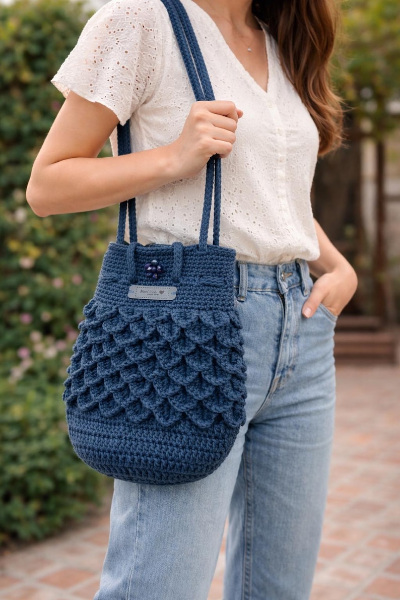 Puede incluir: Bolso de crochet azul marino hecho a mano con un patr&oacute;n &uacute;nico en forma de escamas. El bolso tiene un cierre de cord&oacute;n y correas resistentes. Una peque&ntilde;a etiqueta con texto y un bot&oacute;n decorativo a&ntilde;aden detalles.