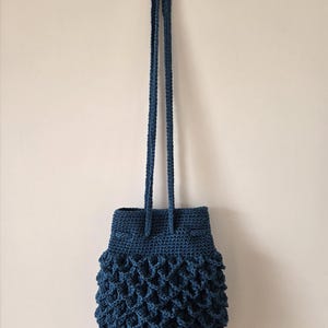 Puede incluir: Un bolso de ganchillo azul oscuro hecho a mano con cierre de cord&oacute;n y correas largas trenzadas. El bolso presenta un patr&oacute;n texturizado en forma de escamas en la parte inferior, agregando un elemento de dise&ntilde;o &uacute;nico. El bolso est&aacute; suspendido contra un fondo neutral.
