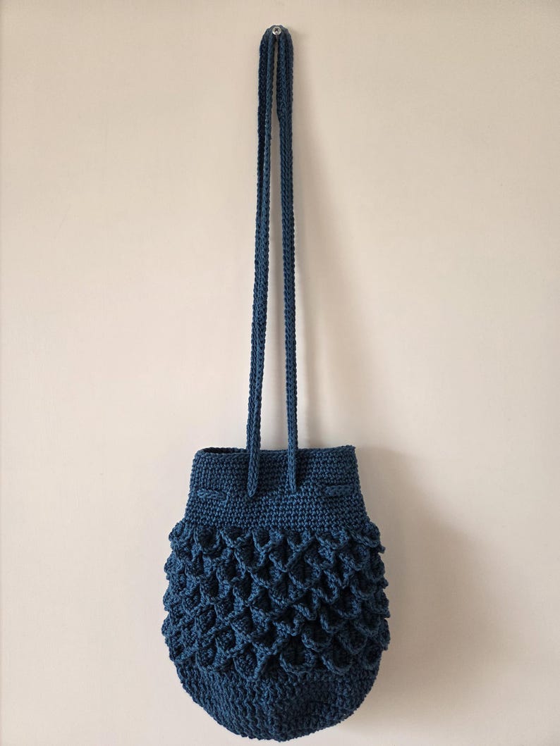 Puede incluir: Un bolso de ganchillo azul oscuro hecho a mano con cierre de cord&oacute;n y correas largas trenzadas. El bolso presenta un patr&oacute;n texturizado en forma de escamas en la parte inferior, agregando un elemento de dise&ntilde;o &uacute;nico. El bolso est&aacute; suspendido contra un fondo neutral.