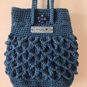Puede incluir: Bolso azul hecho a mano con ganchillo, con un patr&oacute;n de escamas en la mitad inferior. El bolso tiene una etiqueta que dice "Hand Made with love" y una decoraci&oacute;n de cuentas. El bolso tiene dos asas trenzadas.