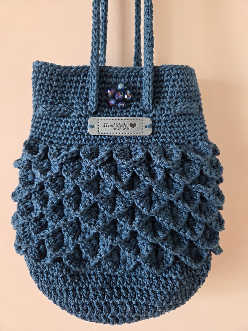 Puede incluir: Bolso azul hecho a mano con ganchillo, con un patr&oacute;n de escamas en la mitad inferior. El bolso tiene una etiqueta que dice "Hand Made with love" y una decoraci&oacute;n de cuentas. El bolso tiene dos asas trenzadas.