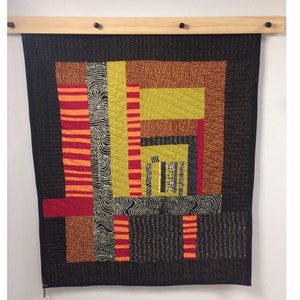 May include: A modern quilt with a geometric design, featuring blocks of red, orange, yellow-green, and black fabrics. The quilt is mounted on a wooden rod with black pegs. The quilt's dimensions are not visible.