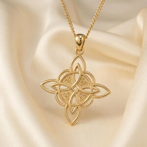 May include: A gold-colored pendant necklace featuring a Celtic knot design. The pendant is a four-pointed shape with intricate interwoven patterns. The necklace hangs on a delicate gold chain, set against a soft, cream-colored background.