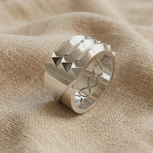 Könnte beinhalten: Ein breiter silberfarbener Ring mit polierter Oberfläche und einer Reihe erhabener pyramidenförmiger Nieten am oberen Rand. Das Band hat ein dickes Profil und ein durchbrochenes Design an der Seite mit einem sich wiederholenden Unendlichkeitsmuster.
