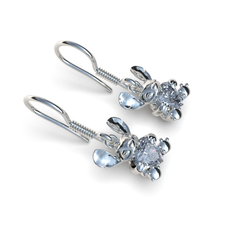 May include: A pair of silver-colored earrings featuring a cartoon character design. Each earring has a hook, a coiled spring, and a character figure with a large, clear gemstone center. The earrings are set against a white background.