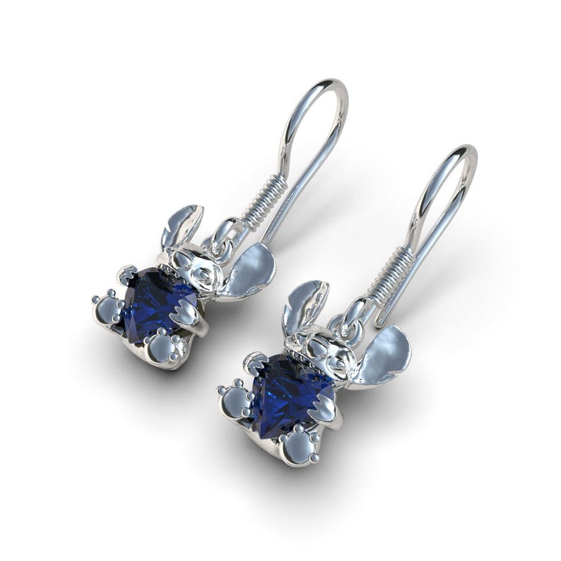 May include: A pair of silver-toned earrings featuring a cartoon character design. Each earring has a blue gemstone at the center of the character's body. The earrings have a hook style closure.